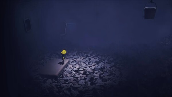 Screenshot from Little Nightmares showing a small figure in a yellow raincoat standing on a mattress amidst scattered shoes in a dark room.